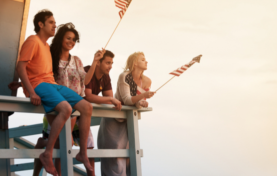 Four friends sitting on lifeguard stand at beach waving American flags