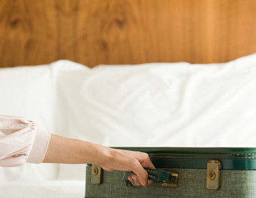 a woman putting a green suitcase on top of a white linen bed