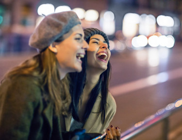 two women standing next to a street laughing with lights blurred in the background
