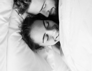 black and white image of a man and woman laying underneath white bed sheets