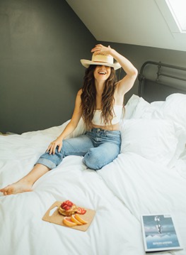 a woman wearing a wide brim hat sitting on a white linen bed with a tray of fruit on the bed