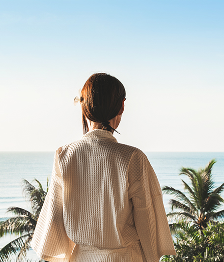 a woman in a bathrobe looking at a view of the beach with palm trees