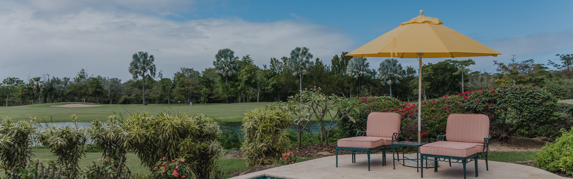 Two light pink cushioned chairs under a yellow umbrella with a gold course behind it,
