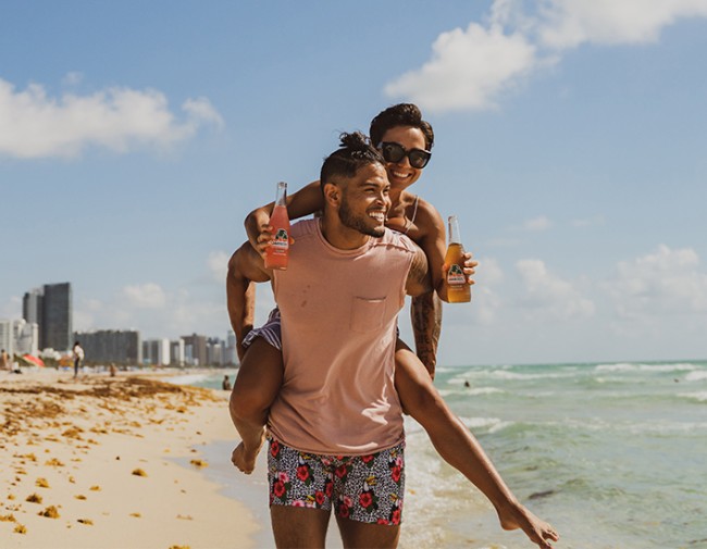 girl on the piggy-backing on a guy as he walks down the beach and she holds onto two jarritos