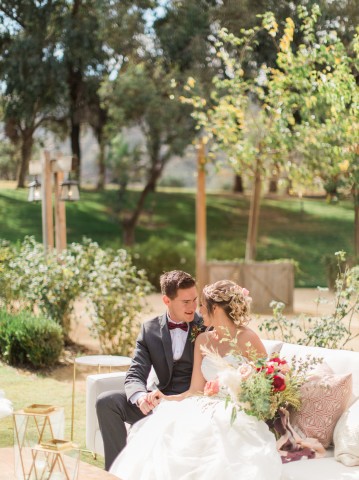 Bride and groom holding hands on couch with greenery in the background