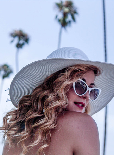 
                            blonde woman with curly hair wearing white sunglasses and a white sunhat looking back 