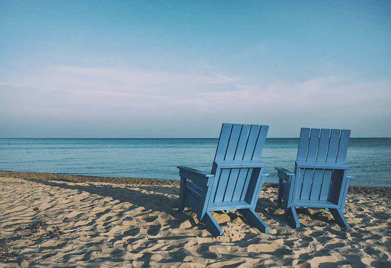 two blue wooden beach chairs in the sand while facing the ocean