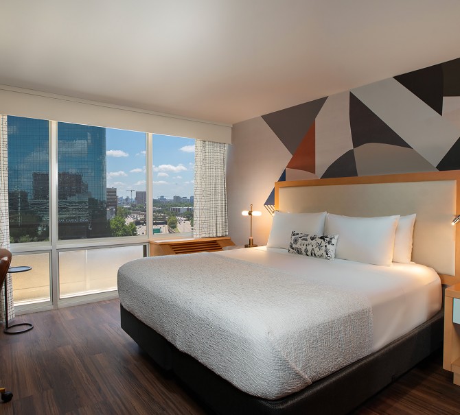 suite with king bed and windows overlooking the city with wooden side table and abstract wallpaper behind bed