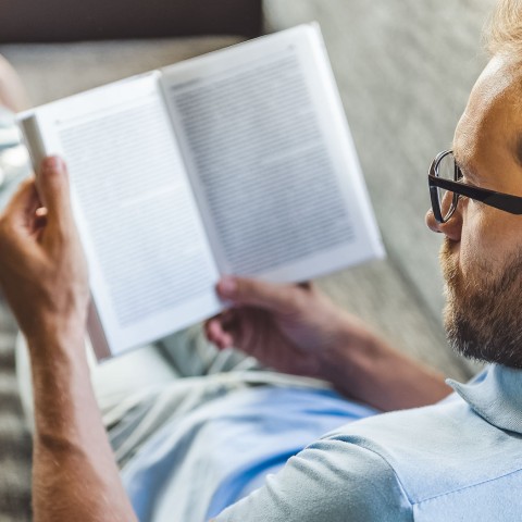 man sitting on bed reading a book with glass on