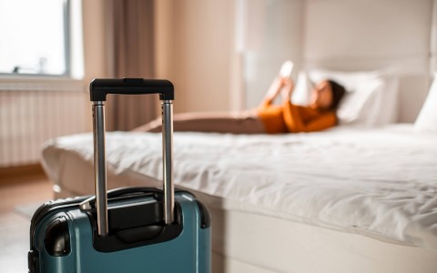 blue suitcase at the foot of the bed and a woman on her phone laying in bed