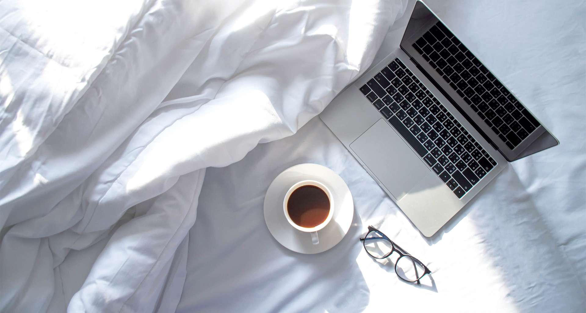 overhead view of a laptop, cup of coffee, and glasses on a bed