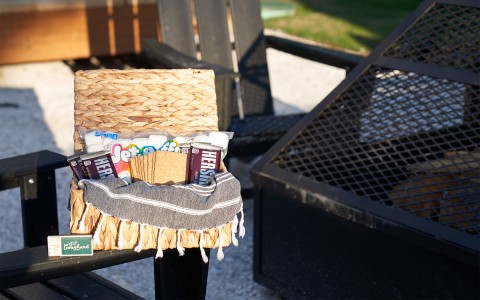 A wicker basket filled with supplies to make smores next to a fire pit and the sun setting.