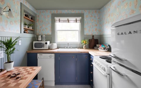 A small kitchen with white appliances and blue cabinets with the sun shining in from the window.