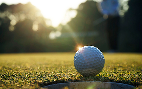 A close up of a golf ball next to the hole and the sun shining in the distance.