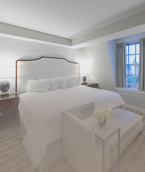 King bed with white sheets, lounger at foot of bed, two nightstands & window
