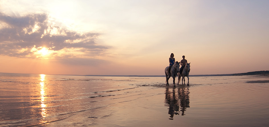 two people riding horses on the beach at sunset