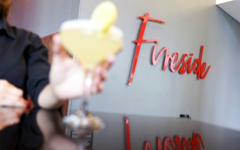 a lady holding a yellow cocktail with the Fireside logo in the background on the wall