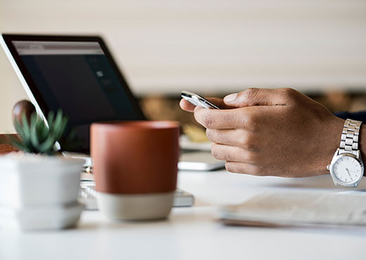 Close up of a man sitting at a desk working on his phone with his computer open on the desk