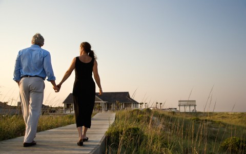 Couple holding hands & walking on wooden path toward building 