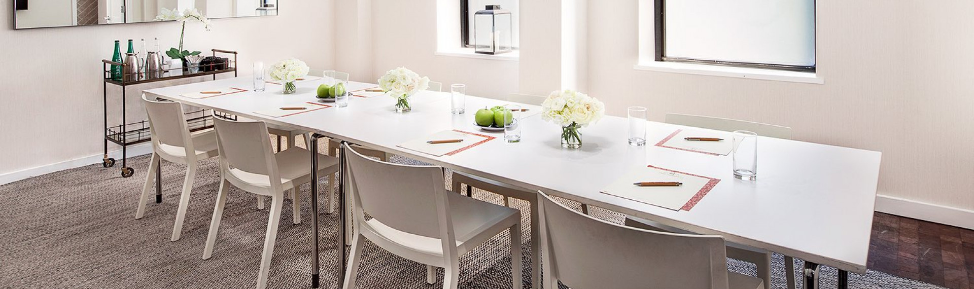 a long white conference table with decorative flowers, apple trays, pens, paper, and empty glasses on the table and a bar cart is against the wall