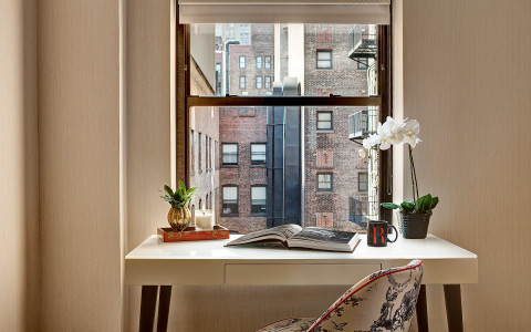 A chair and desk in front of a window looking out to a red brick building. the desk has an open book, plants, and a coffee mug with the redbury logo