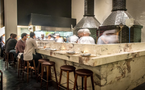 marble bar area at a restaurant with two large black brick ovens and employees walking around behind the counter