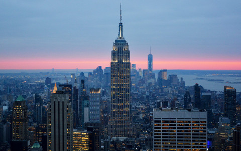 the tall empire state building lit up surrounded by other buildings with a light blue and pink sky in the distance
