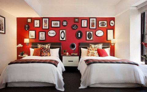 two beds with white sheets, decorative patterned pillows, and a plaid blanket in front of a bright red wall filled with different picture frames