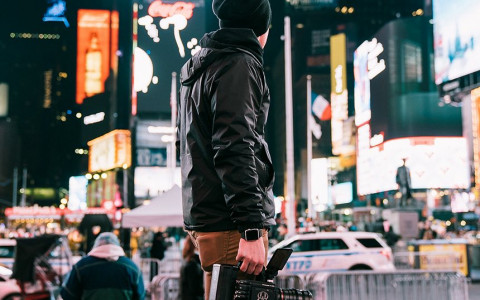 A man wearing a beanie and jacket standing in front of the bright screens at night in times square