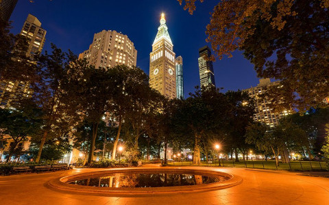 A circular pond in madison square park lit up at night surrounded by trees with a view of buildings lit up in the distance