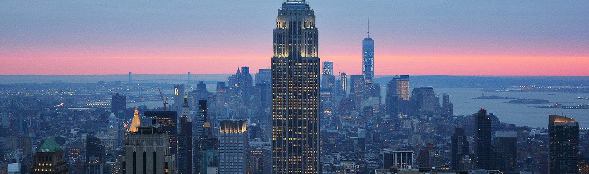 the tall empire state building surrounded by other buildings with a light blue and pink sky in the distance