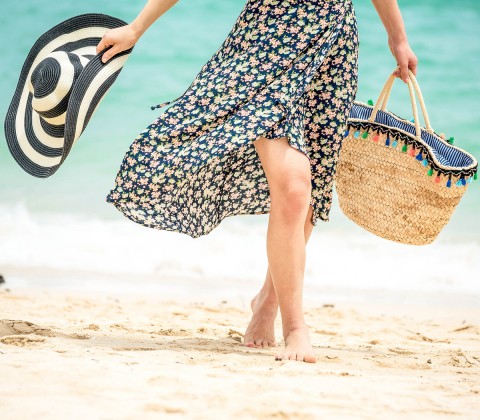 Close up of a lady wearing a flowered dress and carrying a beach hat and a bag