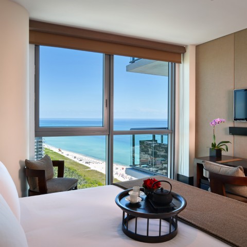 a room with a large bed and desk overlooking the ocean during the day