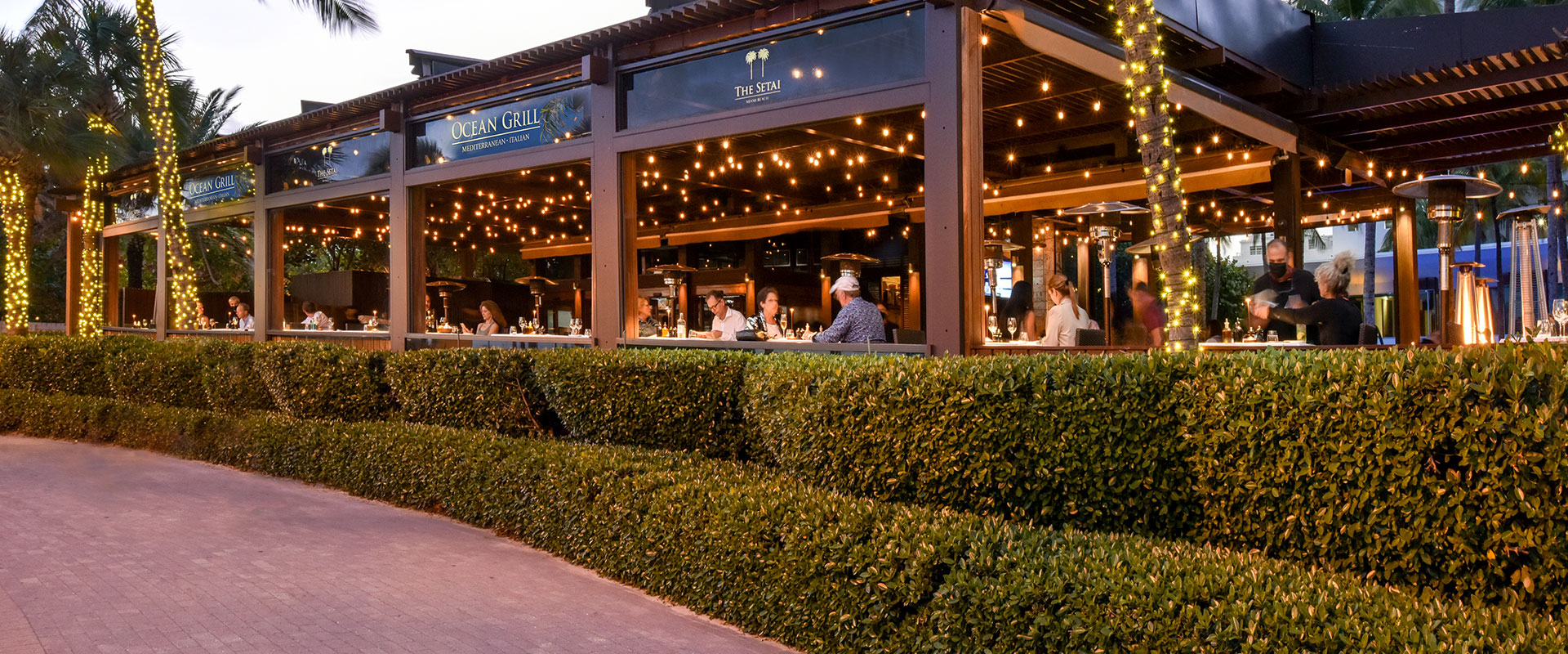 an outdoor area for dining with lots of landscaped bushes in front and lit palm trees at dusk