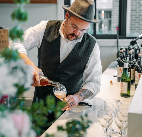 bartender wearing a grey fedora and vest pouring wine