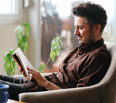 man reading a novel in chair in front of plant