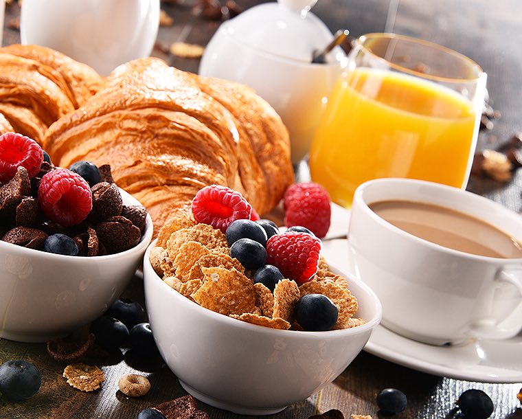 A table full of breakfast foods that includes, bowls of cereal topped with fresh blueberries and raspberries, coffee, croissants, and a glass or orange juice