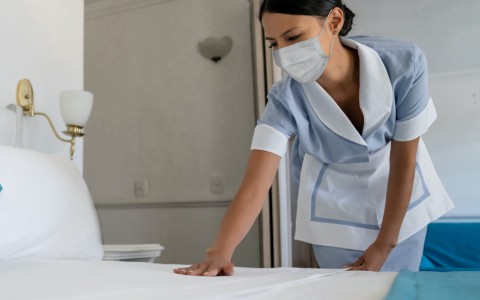 a photo of a hotel maid with a protective mask on making the bed