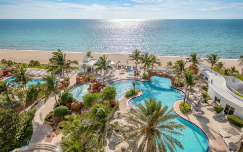 a birds eye view of two pools with a lot of palm trees right on the beach