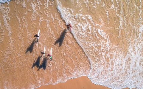 aerial view of four people walking into the ocean with their surfboards 