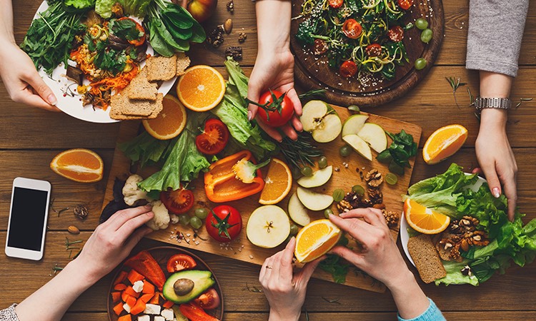 overhead view of a group of friends reaching for fruits and veggies on a wooden serving tray