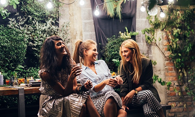 group of three women sitting on a bench at an outdoor restaurant having drinks and laughing