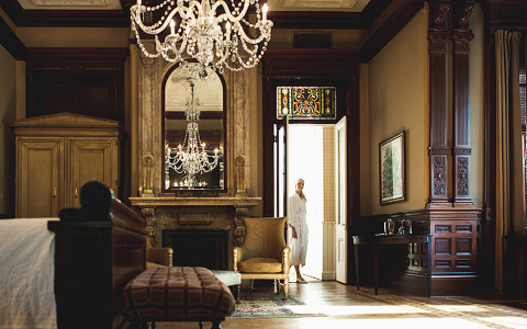 Woman walking into room with king bed, antique wooden accent decor & chandelier