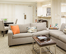 interior of a guest room suite with a gray couch, orange and patterned pillows, and a view of the dining table and kitchen in back