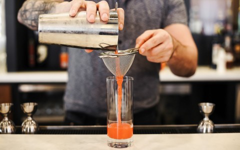 bartender pouring red drink through a strainer into a tall glass