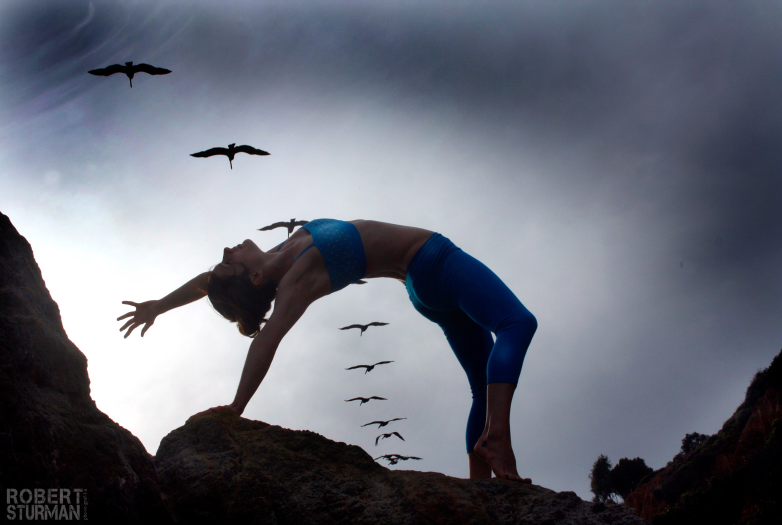 View of a woman doing a yoga pose on a rock and some birds flying at the background