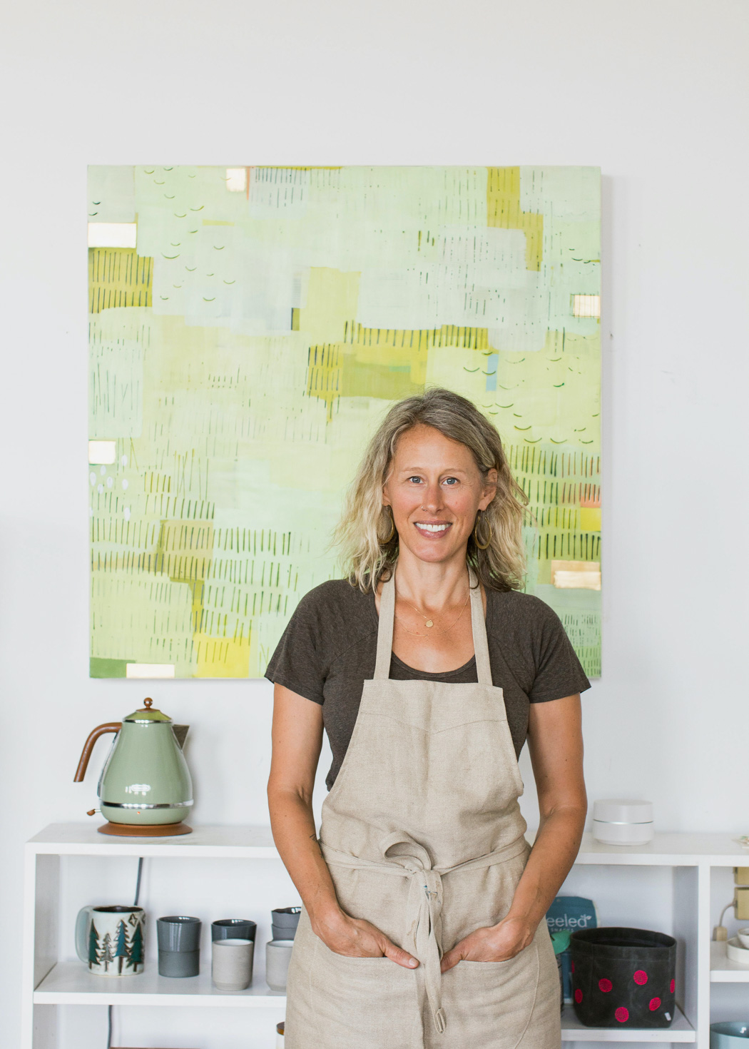  woman smiling wearing a beige apron and a big green paint on the wall at the back