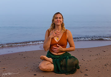a lady sitting on the sand in a yoga pose in front of the beach