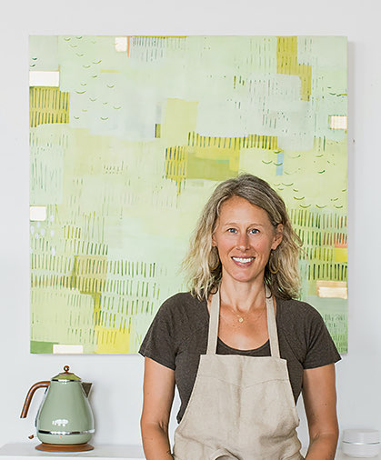  woman smiling wearing a beige apron and a big green paint on the wall at the back