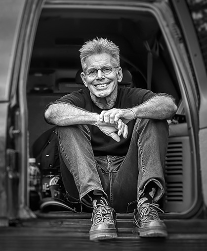 A black and white portrait of a smiling man on sitting on a car